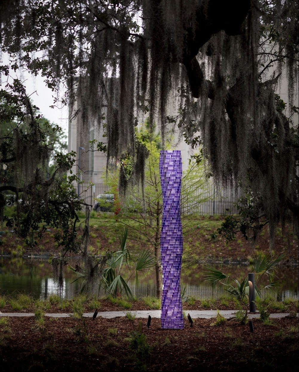 Tall, purple sculpture in a park setting with trees and moss hanging overhead, pathway beside a calm pond.