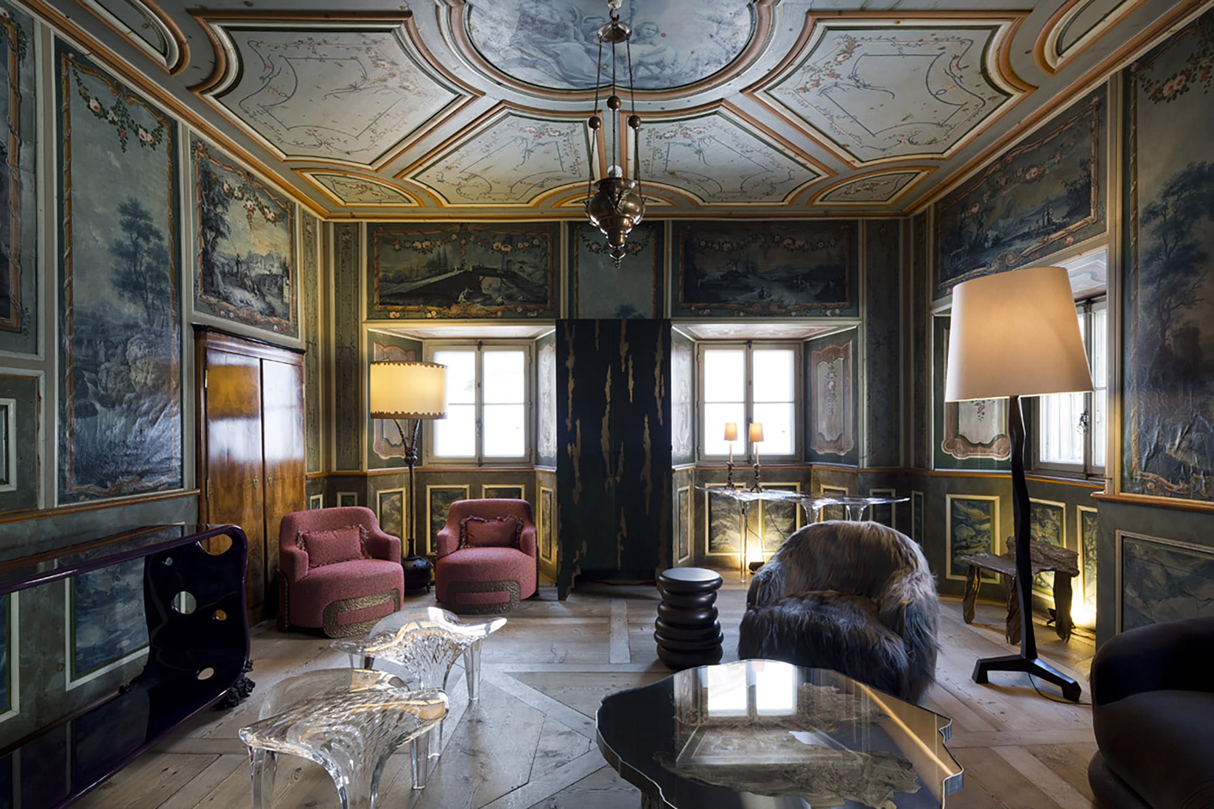Luxurious room with ornate painted walls, vintage armchairs, unique tables, and large lamps, under a decorated ceiling.