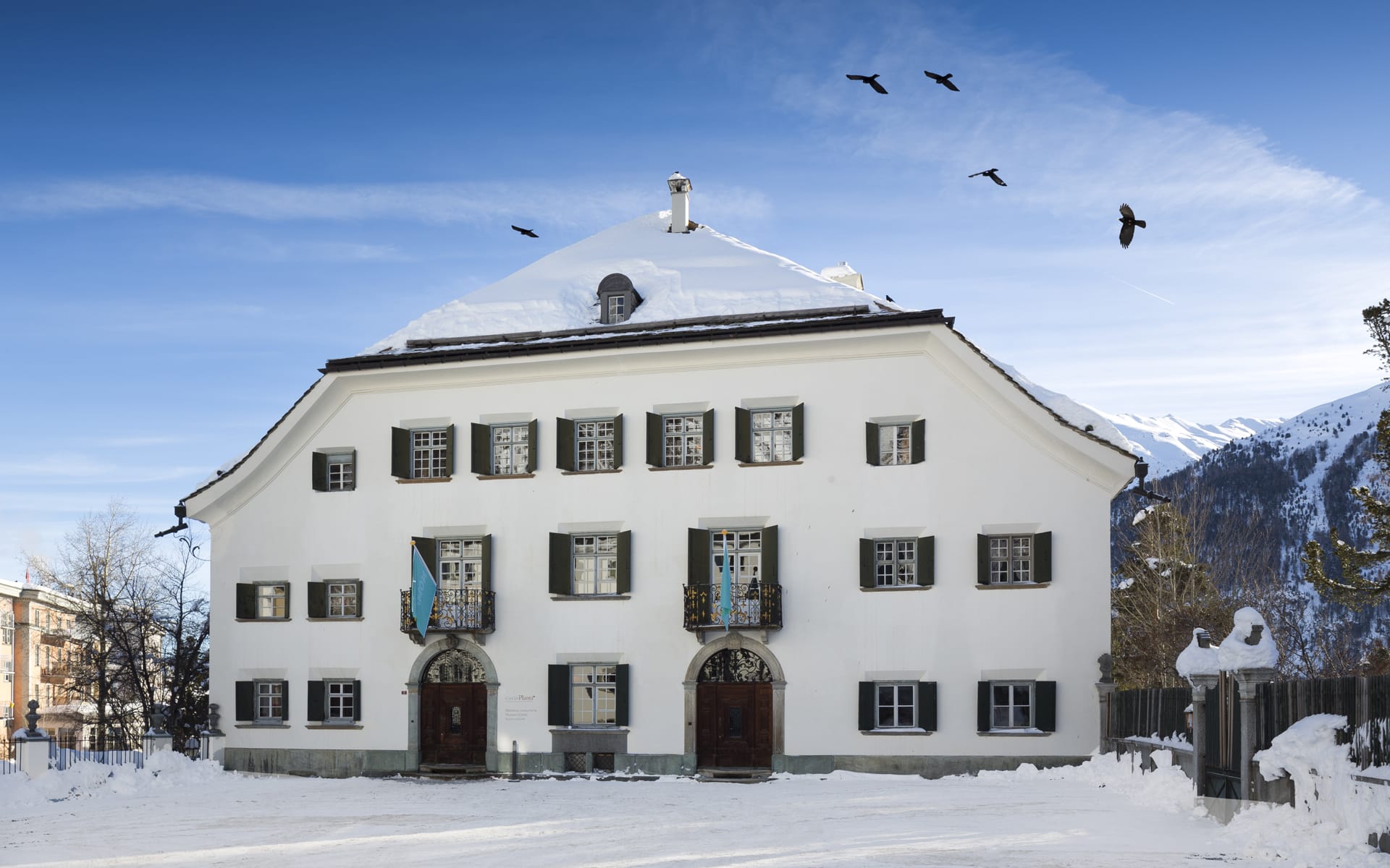 Historic white building with dark shutters in a snowy landscape, birds in the sky, surrounded by snow-covered mountains.