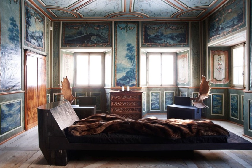 Luxurious room with ornate walls, a rustic bed covered in a dark fur throw, and large antler chair accents.