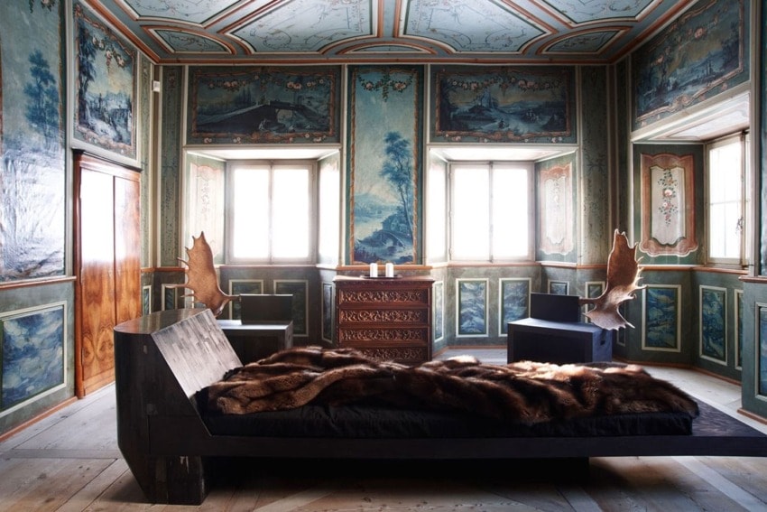 Luxurious room with ornate walls, a rustic bed covered in a dark fur throw, and large antler chair accents.