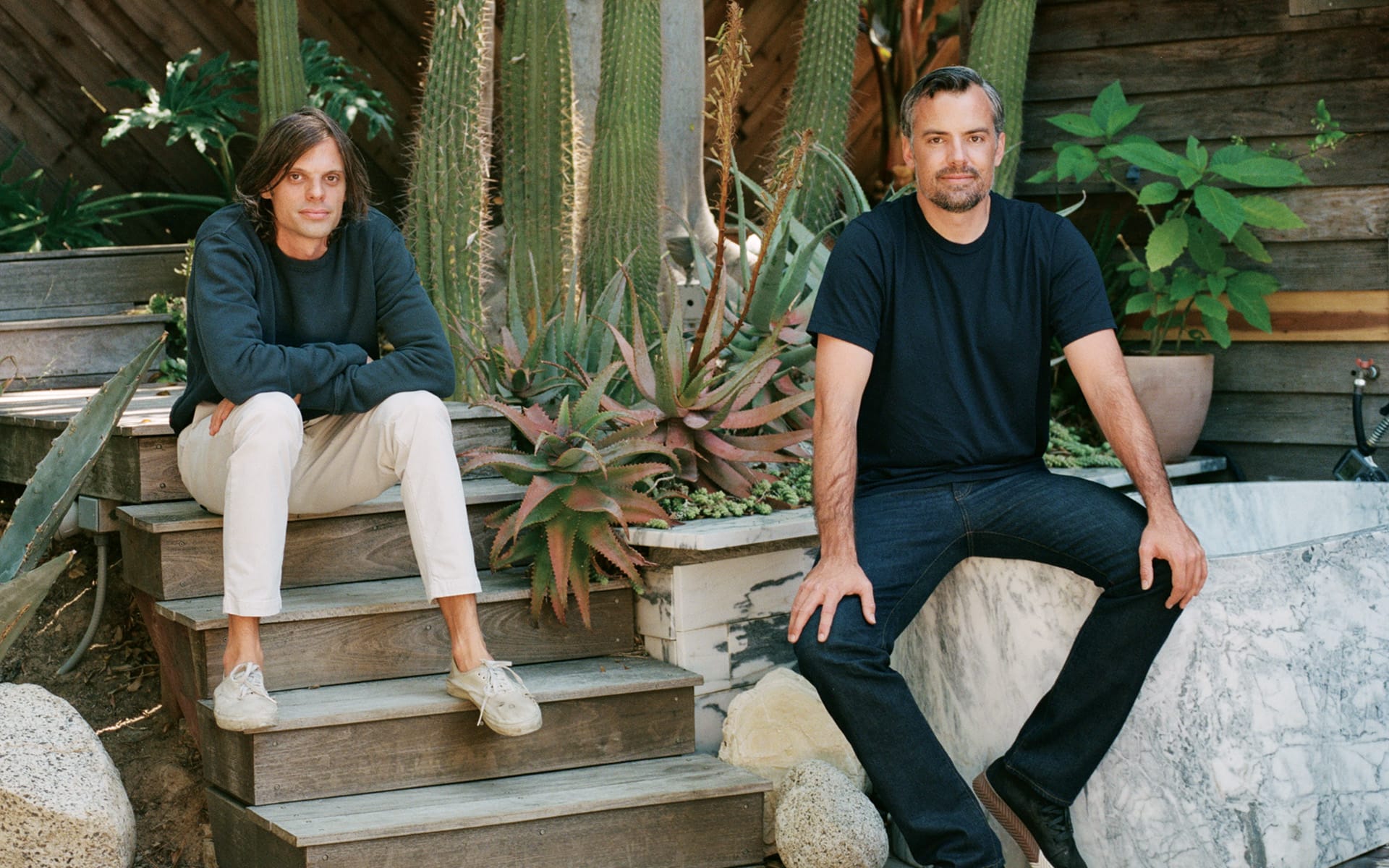 Two men sitting on wooden steps surrounded by plants and greenery in an outdoor setting.