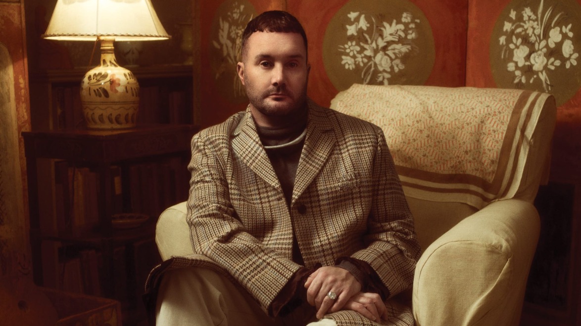 Person sitting in an armchair, wearing a plaid coat, with a lamp and decorative wall in the background.