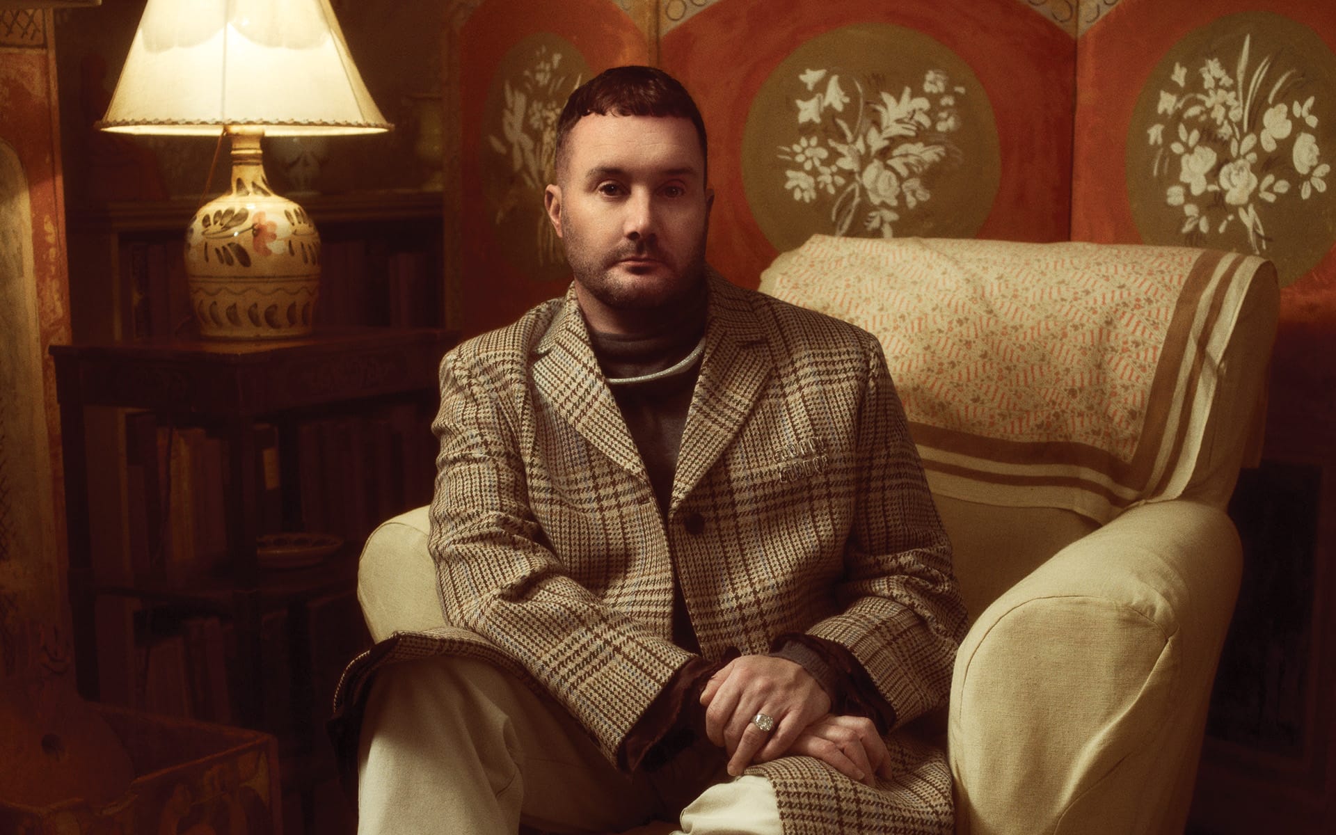 Person sitting in an armchair, wearing a plaid coat, with a lamp and decorative wall in the background.