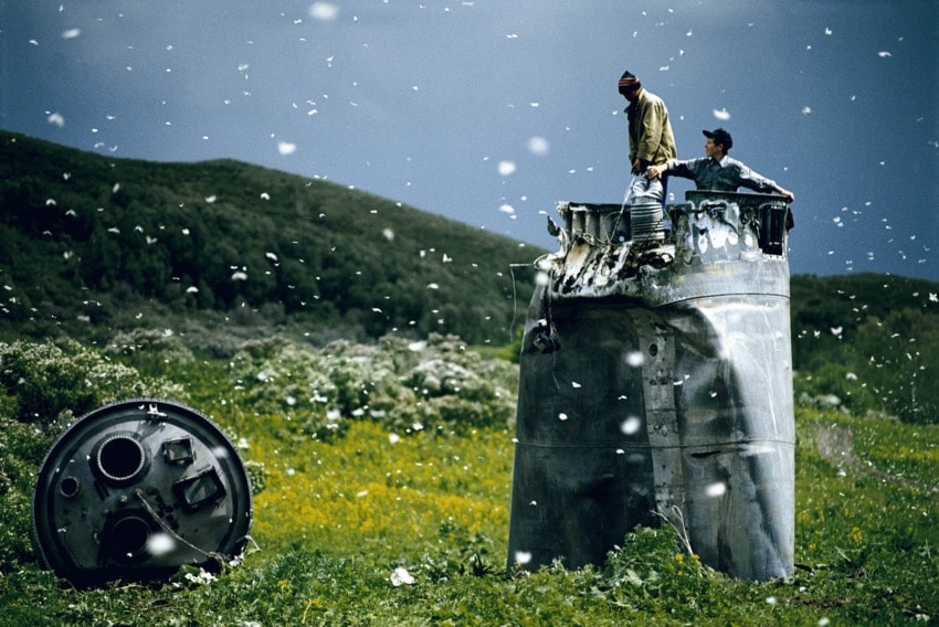 Two men stand atop a large metallic structure in a grassy field with white particles floating in the air.