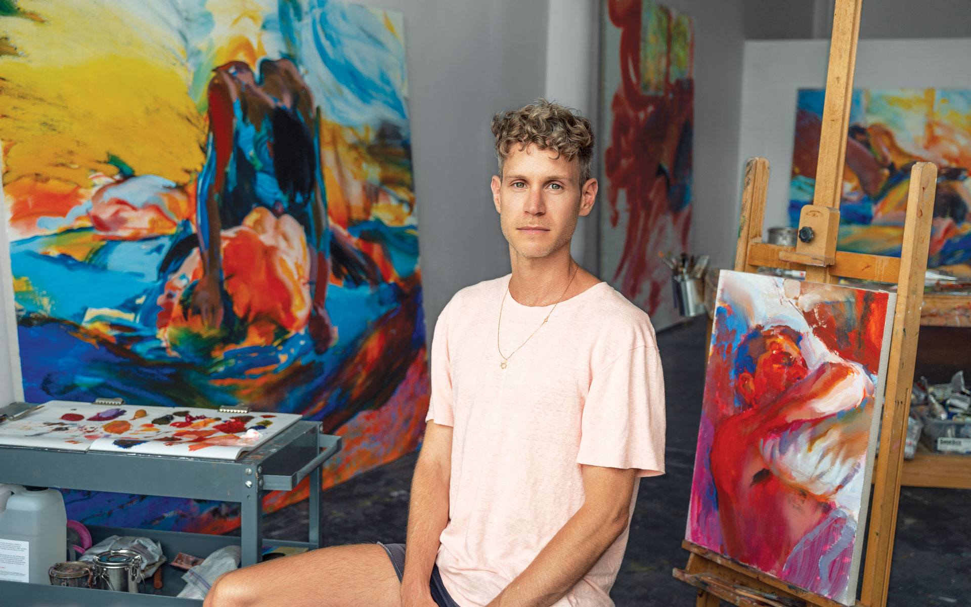 Artist sitting in studio with colorful abstract paintings, wearing a pink shirt, paint supplies and easels visible in the background.