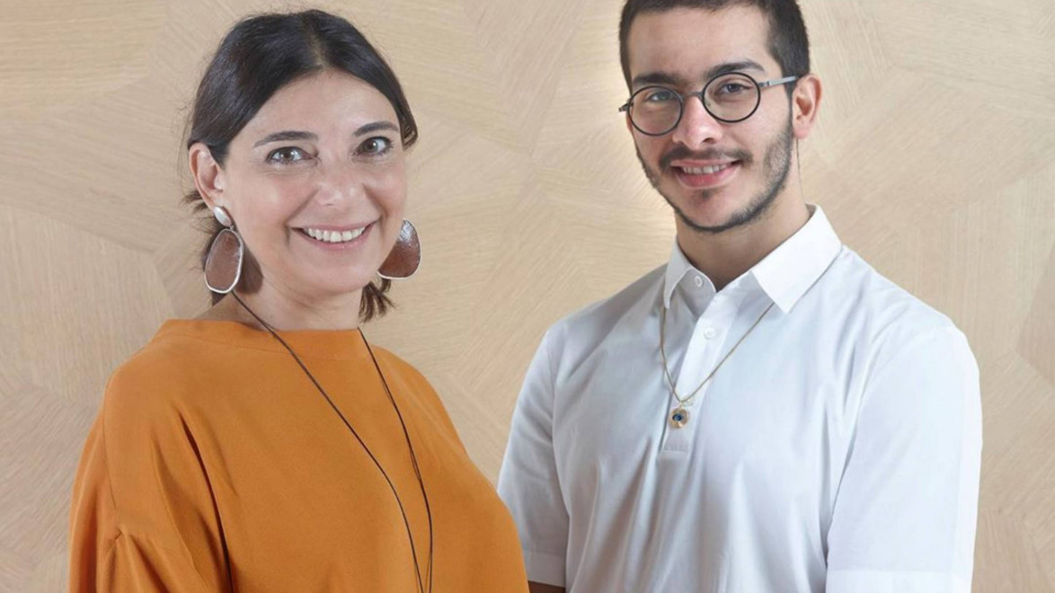 Two people smiling, one wearing an orange top with earrings, and the other in a white shirt with glasses, standing against a light background.