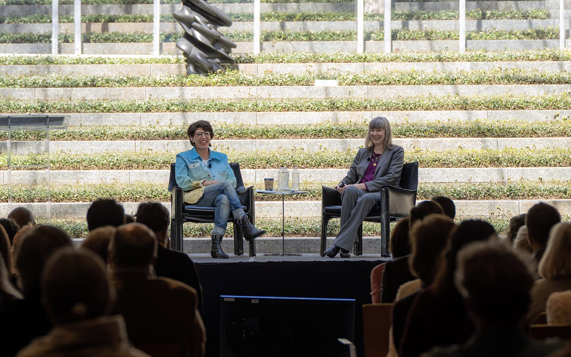 Two people seated and speaking on stage at an outdoor event with an audience in the foreground.