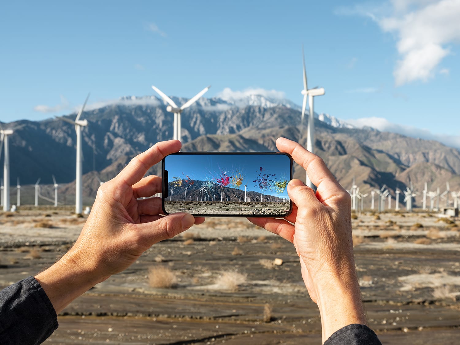 Hands holding a smartphone capturing wind turbines against mountains, with an augmented reality display showing colorful graphics.