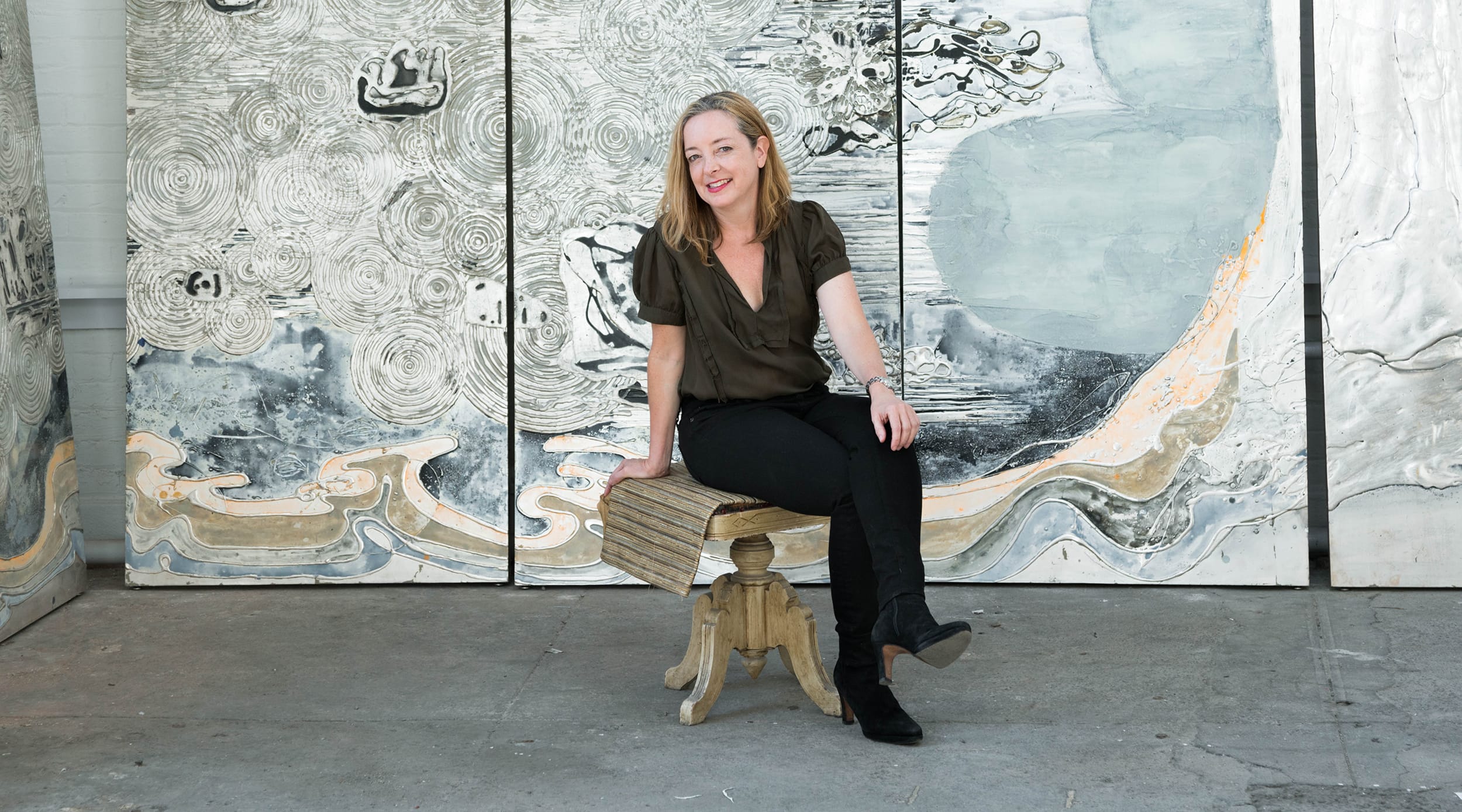 Person sitting on a wooden bench in front of abstract art, wearing a dark shirt and black pants, smiling at the camera.