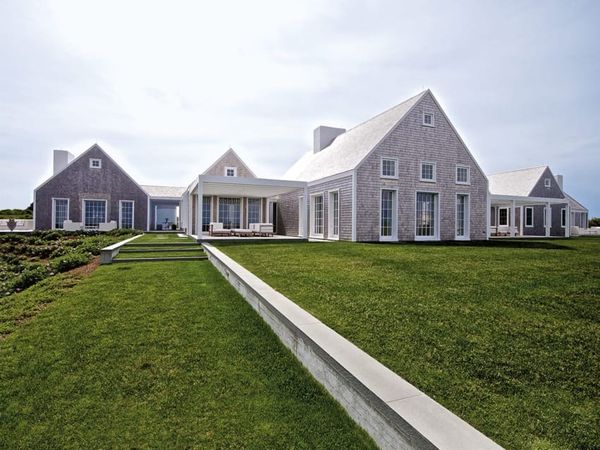 Large, modern coastal home with gray exterior, white trim, expansive lawn, and partly cloudy sky in the background.