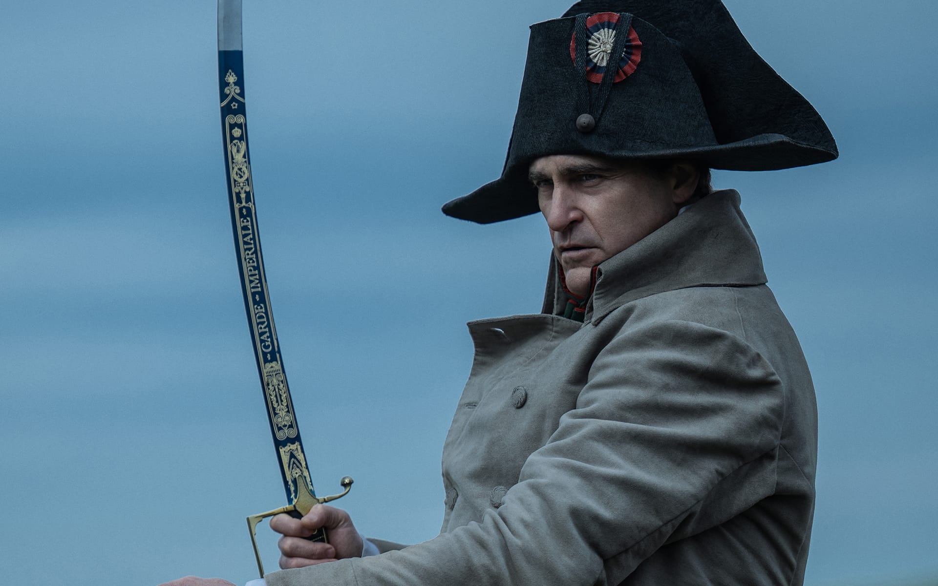 Man in historic military uniform holding a sword with a serious expression against a cloudy sky background.
