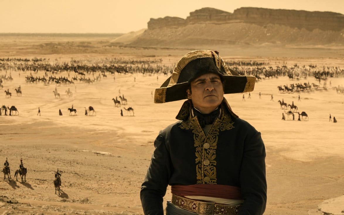 A man in historical military attire stands in a desert, with distant horses and riders against a backdrop of hills.