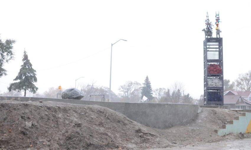 Outdoor art installation with stacked sculptures on a tall structure, surrounded by bare trees and a cloudy sky.