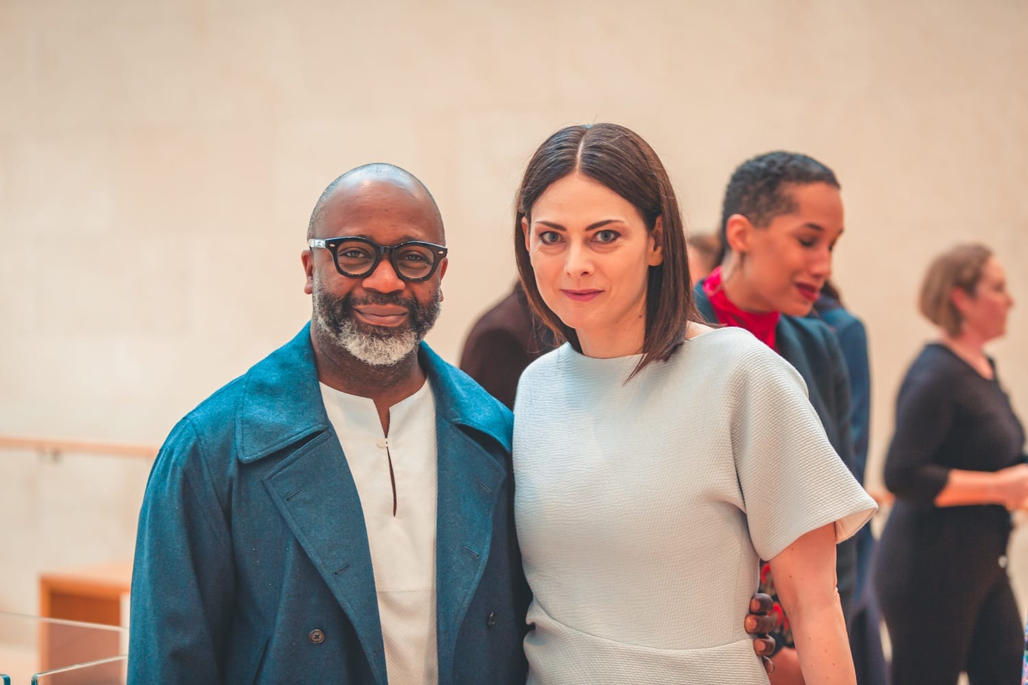 Man in blue coat and woman in white dress smiling at an event with people in the background.