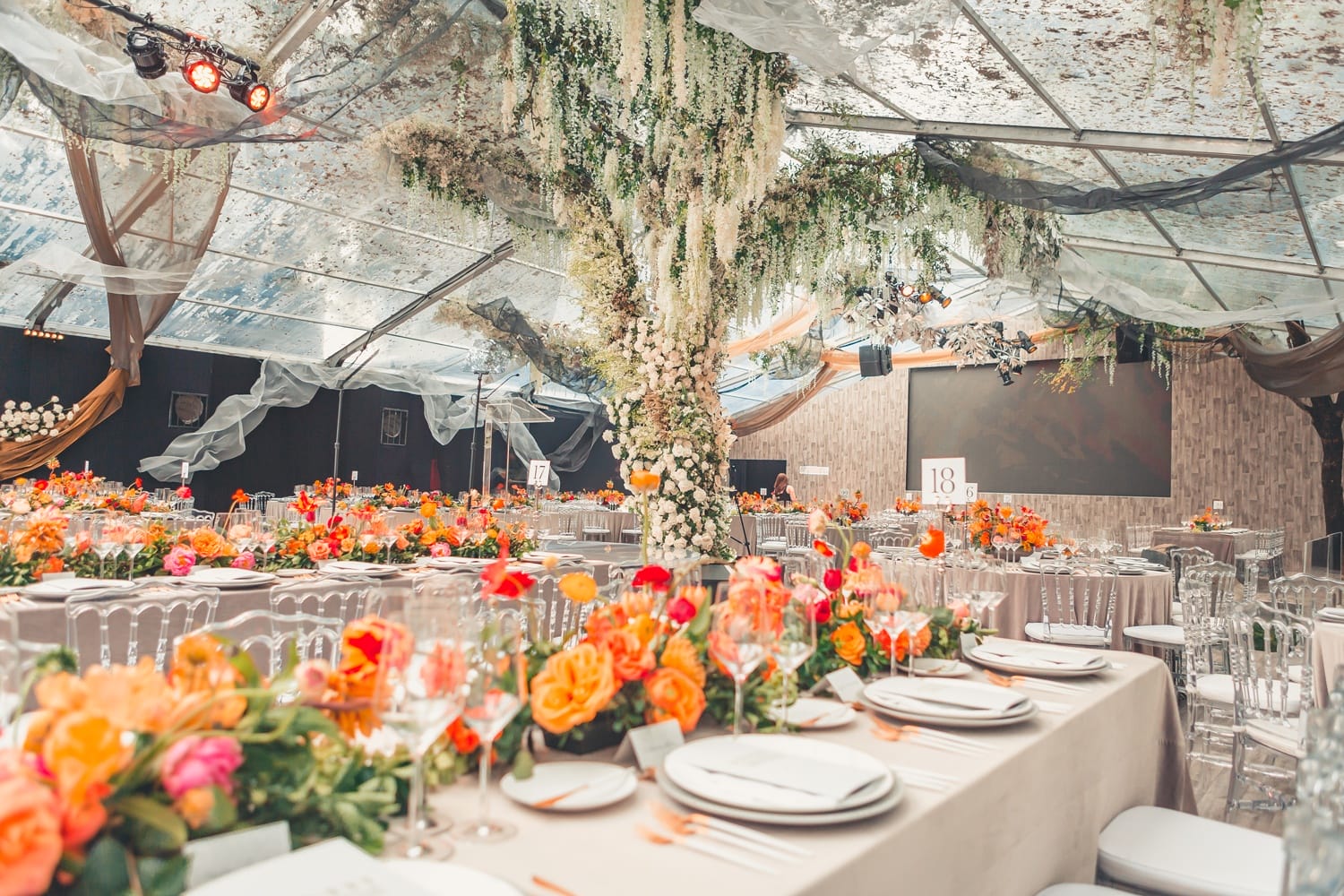 Elegant event setup with tables adorned with vibrant orange and red flowers under a clear canopy with hanging greenery.