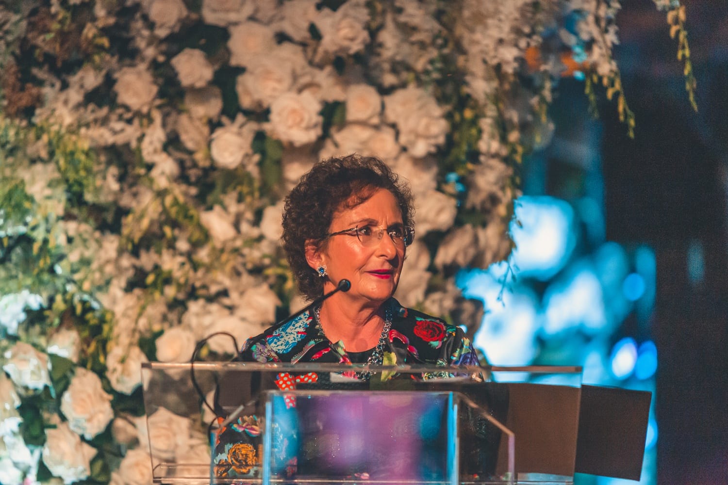 A person speaking at a podium with a floral backdrop, wearing glasses and a colorful floral outfit.