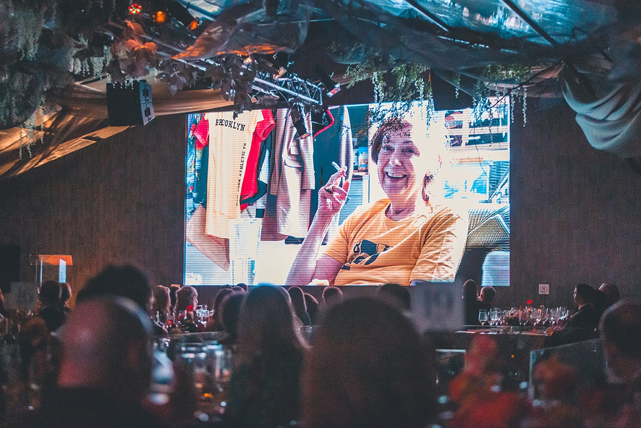 Audience watching a large screen displaying a smiling person during an indoor event.