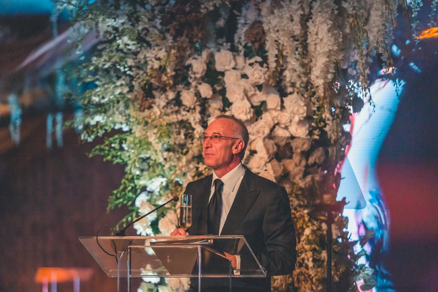 Man speaking at a podium in front of a floral arrangement, during an indoor event.