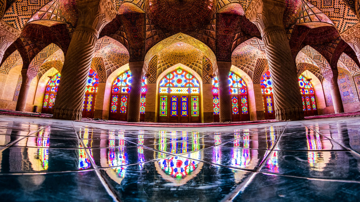 Interior of a mosque with colorful stained glass windows, ornate arches, and reflections on a polished floor.