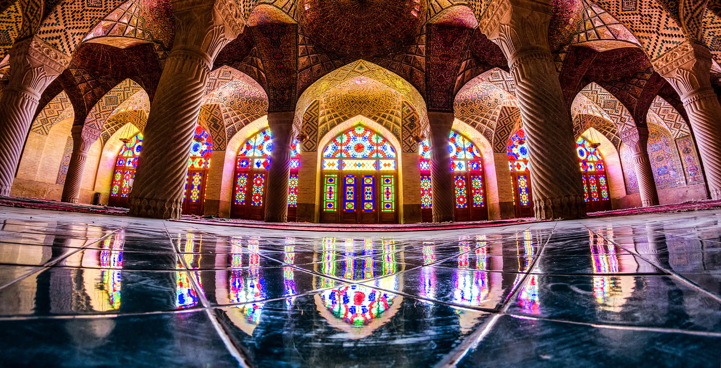 Interior of a mosque with colorful stained glass windows, ornate arches, and reflections on a polished floor.