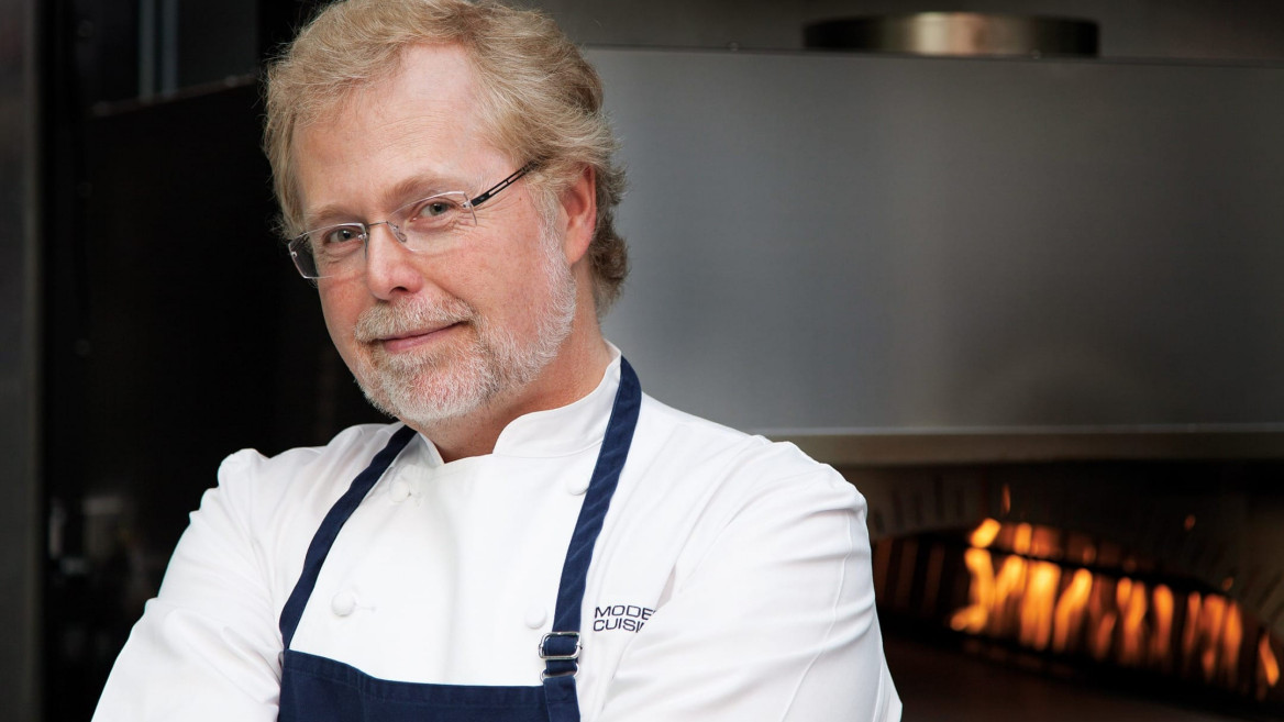Chef in a white coat and blue apron standing confidently in front of an industrial kitchen oven with flames in the background.