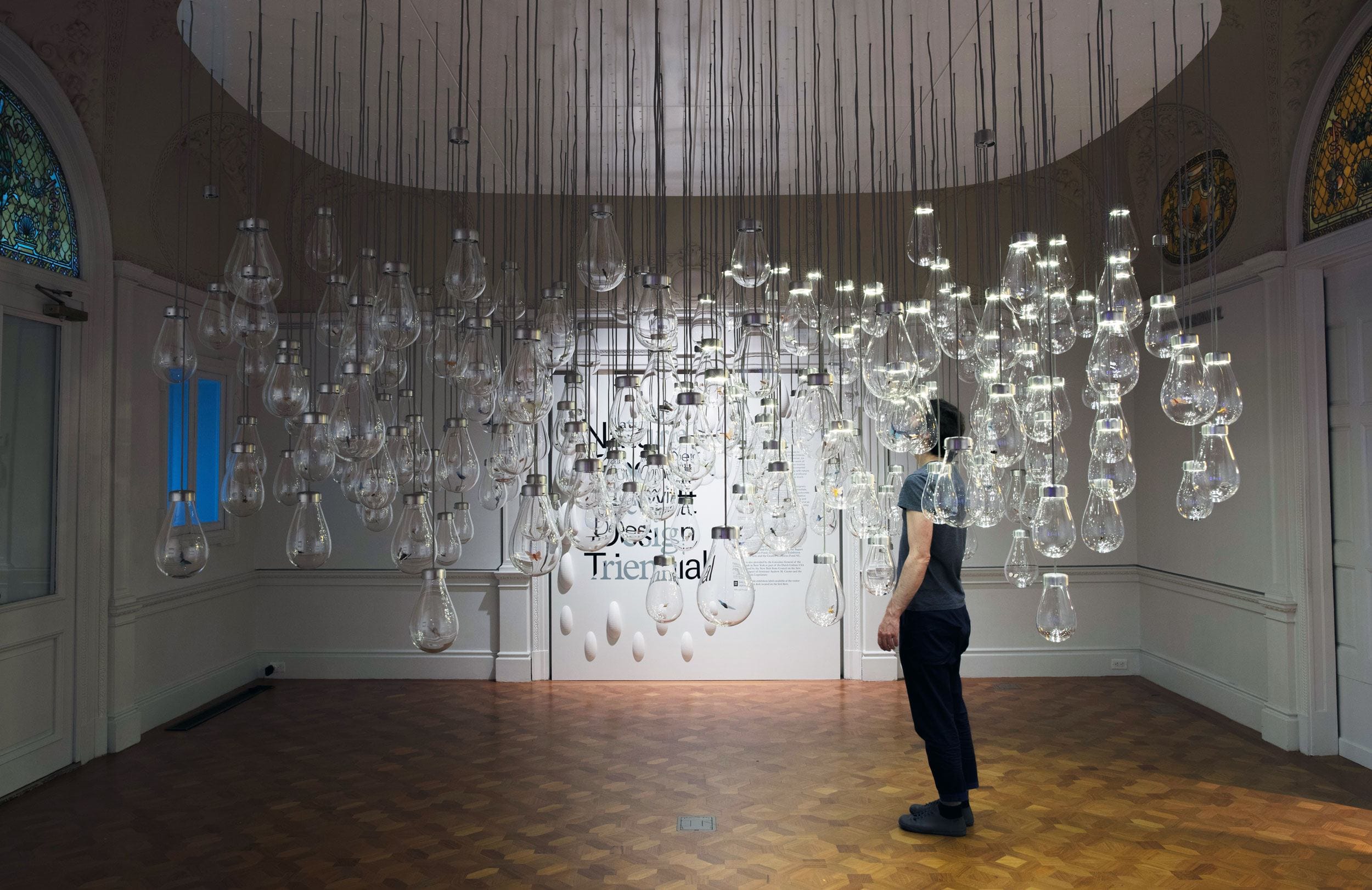 Person observing hanging glass light bulbs in a dimly lit room at a design triennial exhibition.