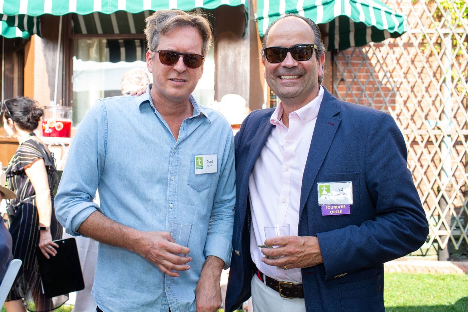 Two men wearing sunglasses pose together outside at an event, holding drinks, with a striped green canopy in the background.