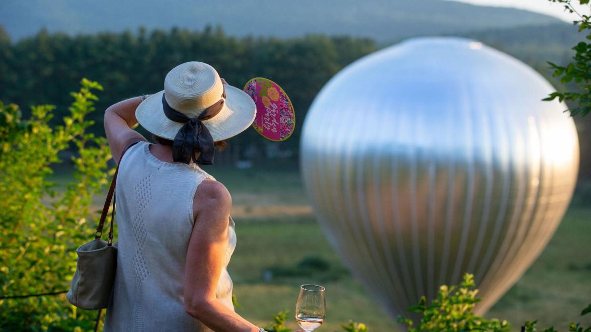 Woman with hat and fan holding a wine glass watches a large shiny balloon in a scenic outdoor setting.