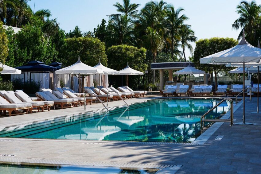 Outdoor pool area with lounge chairs and umbrellas surrounded by palm trees on a sunny day.