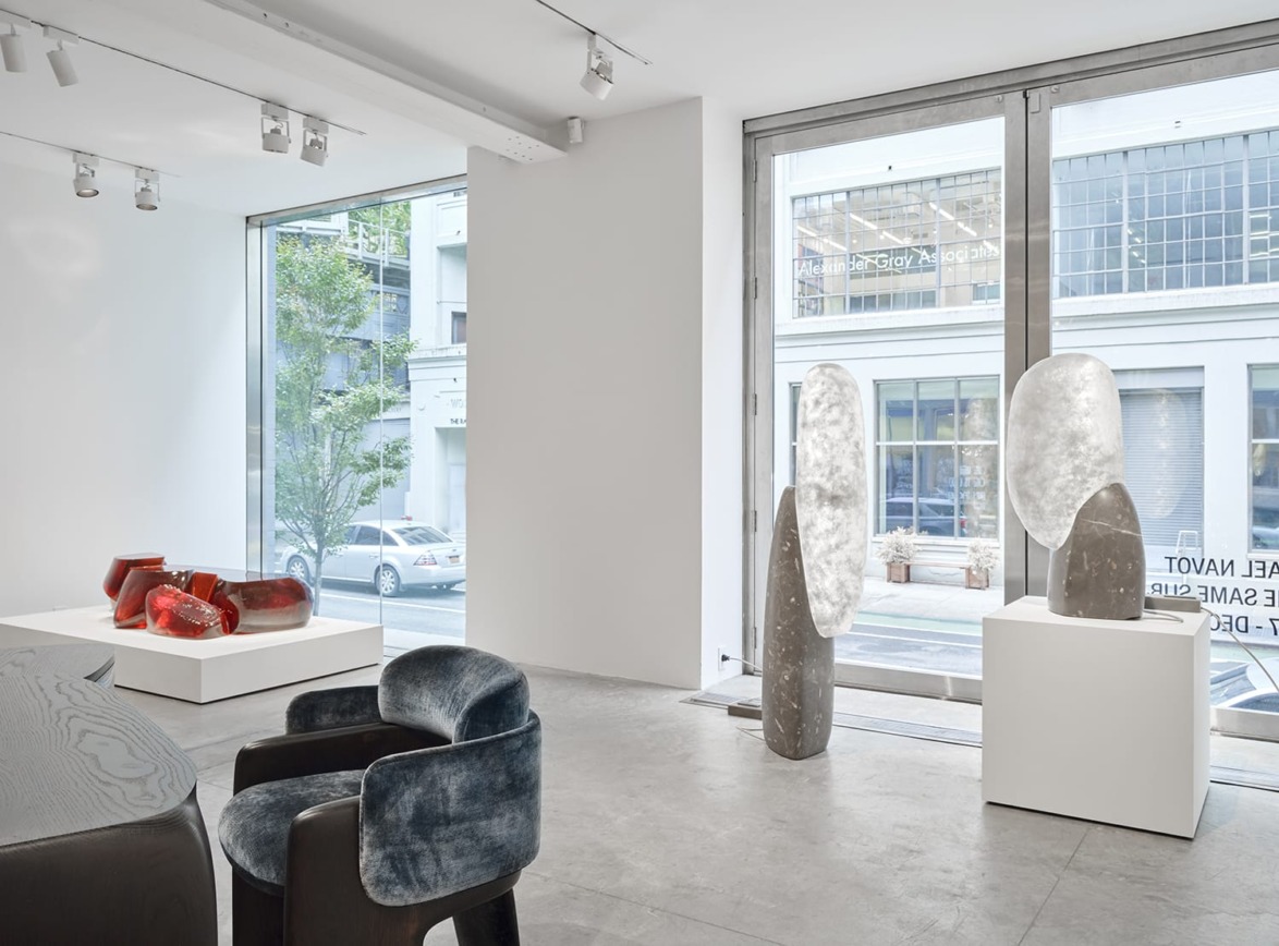 Contemporary art gallery interior with modern sculptures, large windows, natural light, and city street view outside.