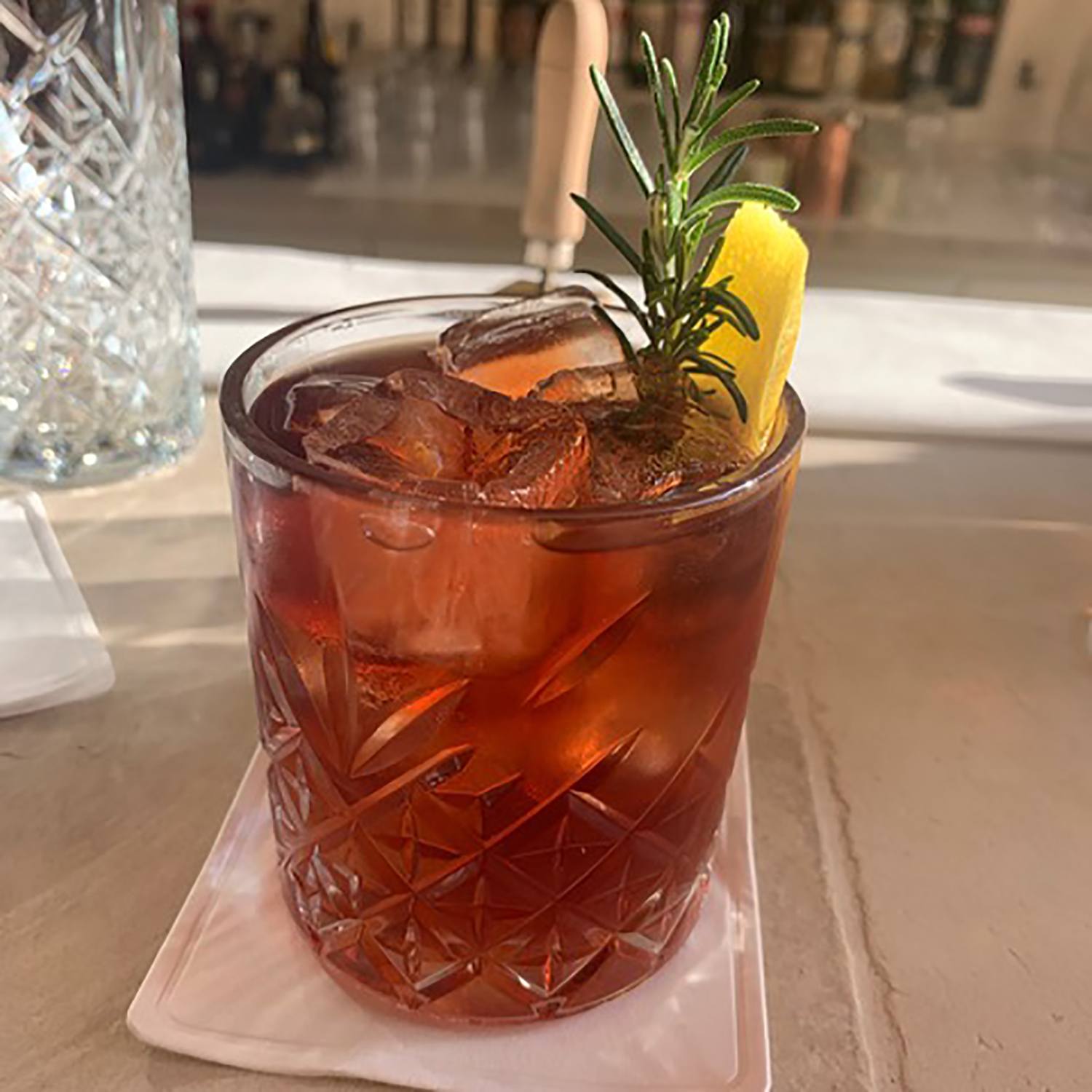 Cocktail with ice and rosemary garnish in a textured glass on a white napkin, bar background with bottles.