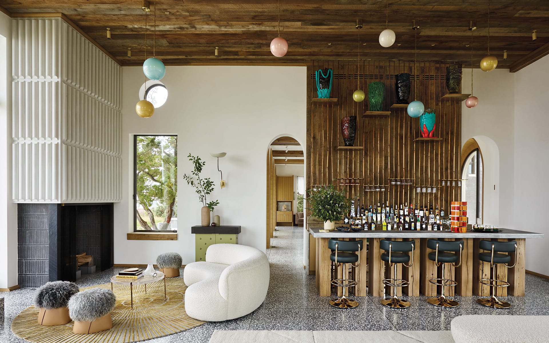 Spacious living room with modern decor, plush seating, a bar with stools, colorful hanging lights, and a large window.