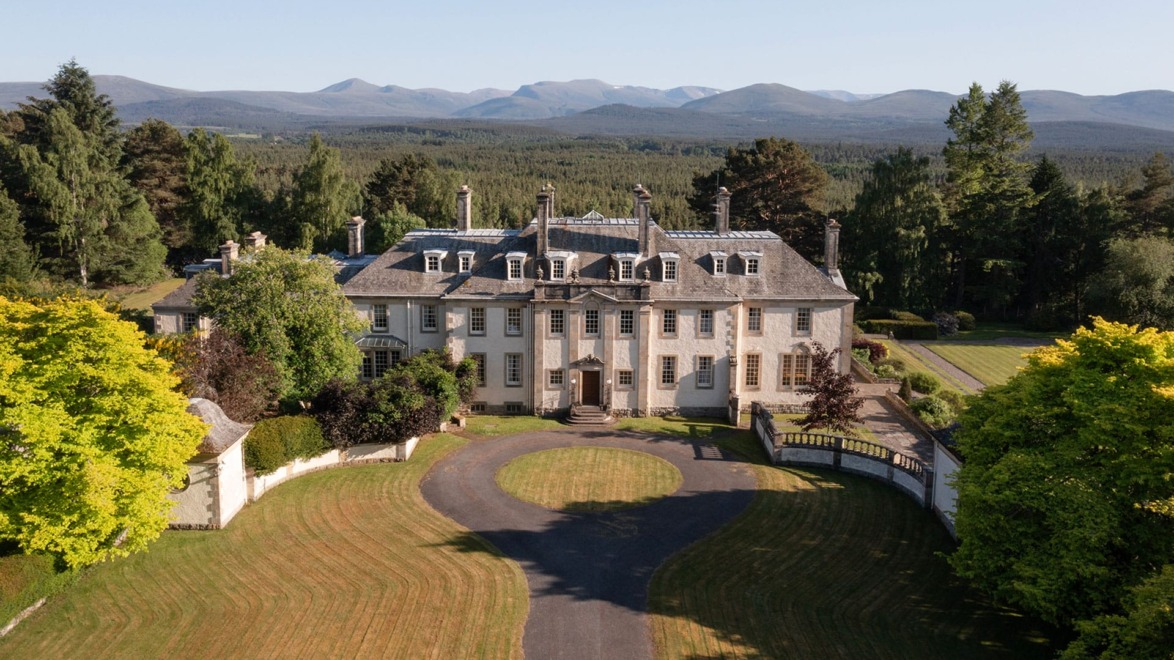 Large historic mansion with multiple chimneys surrounded by lush greenery and mountains in the background.