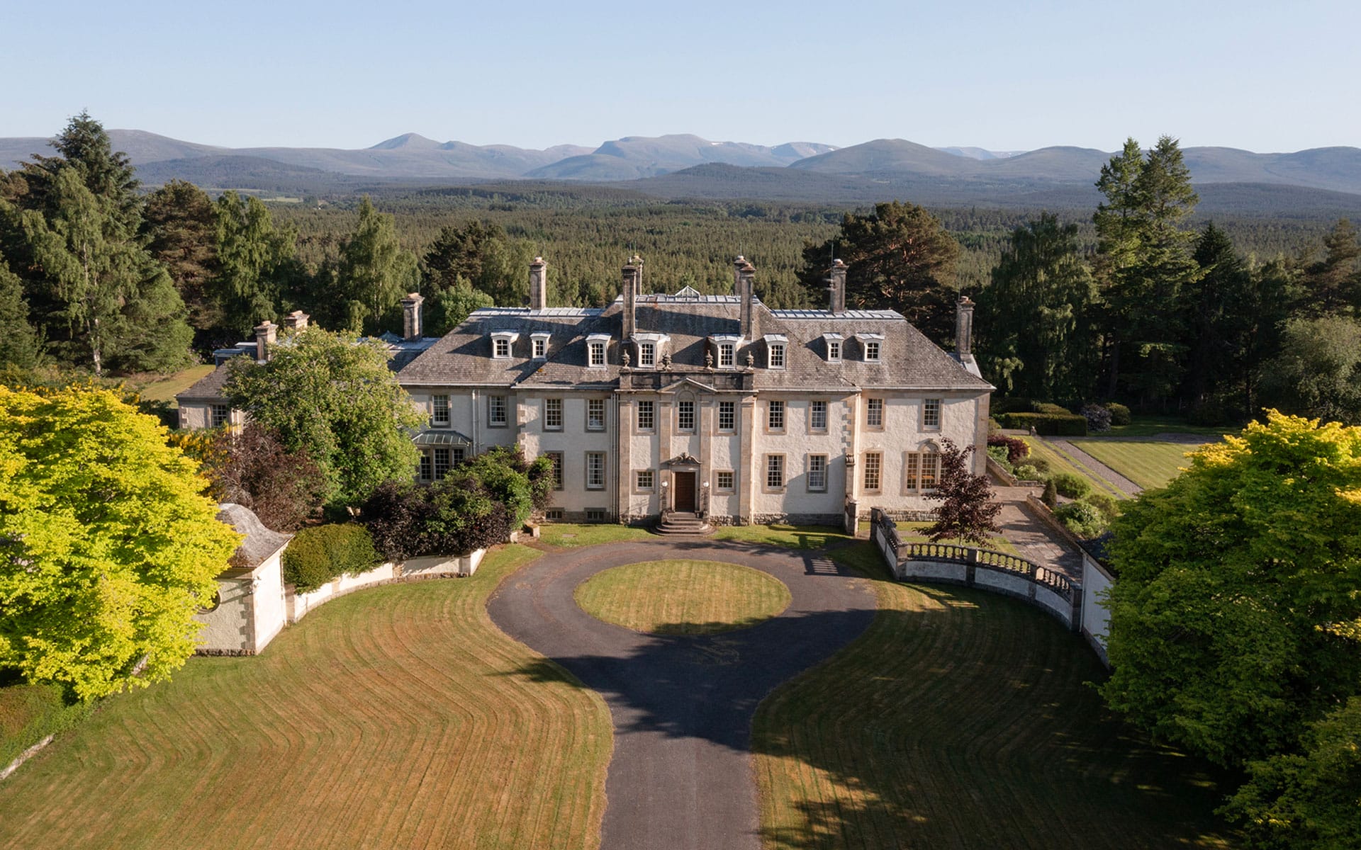 Large historic mansion with multiple chimneys surrounded by lush greenery and mountains in the background.