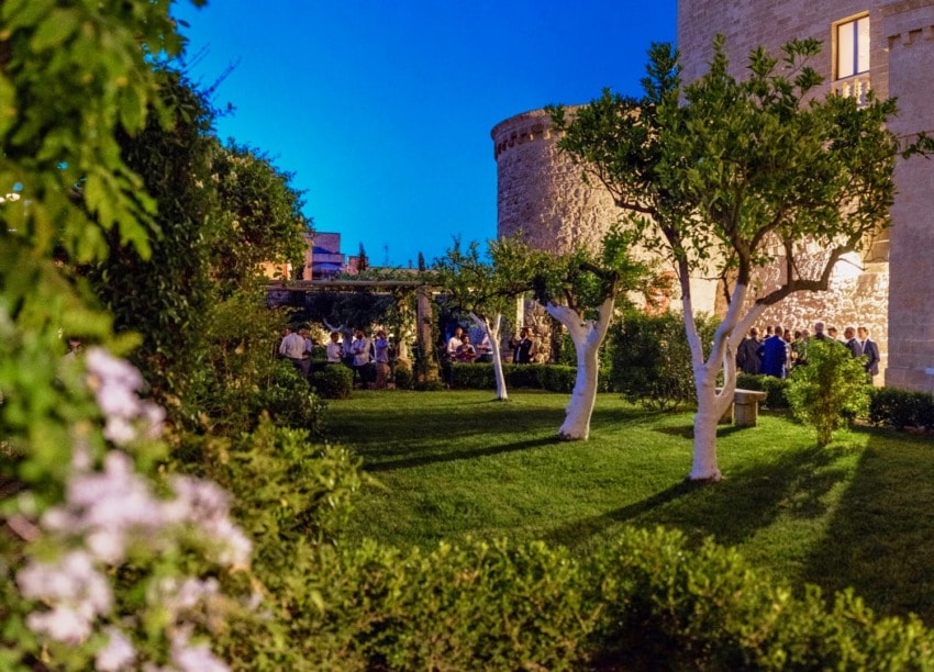 Evening garden party at a historic stone building with guests mingling under trees and vibrant greenery.