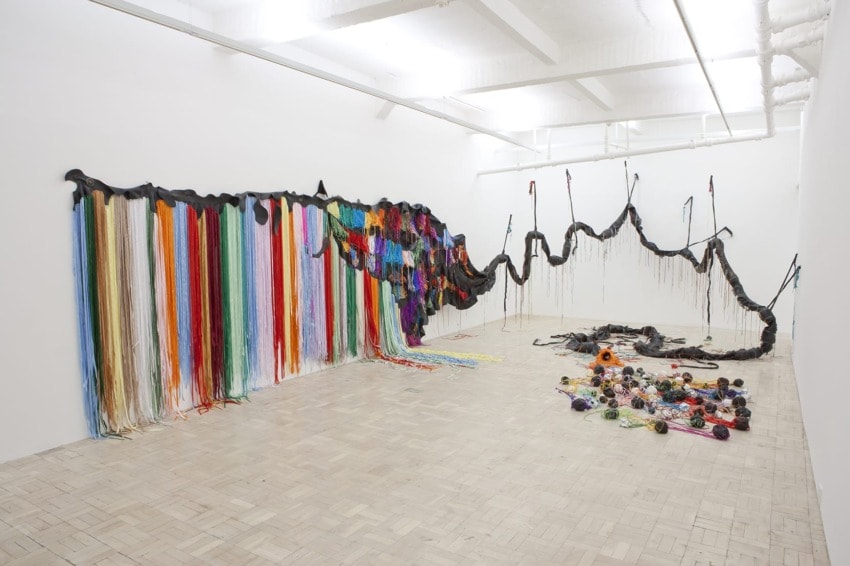 Colorful yarn art installation in a white gallery space with strands and tangled shapes extending across the floor and wall.