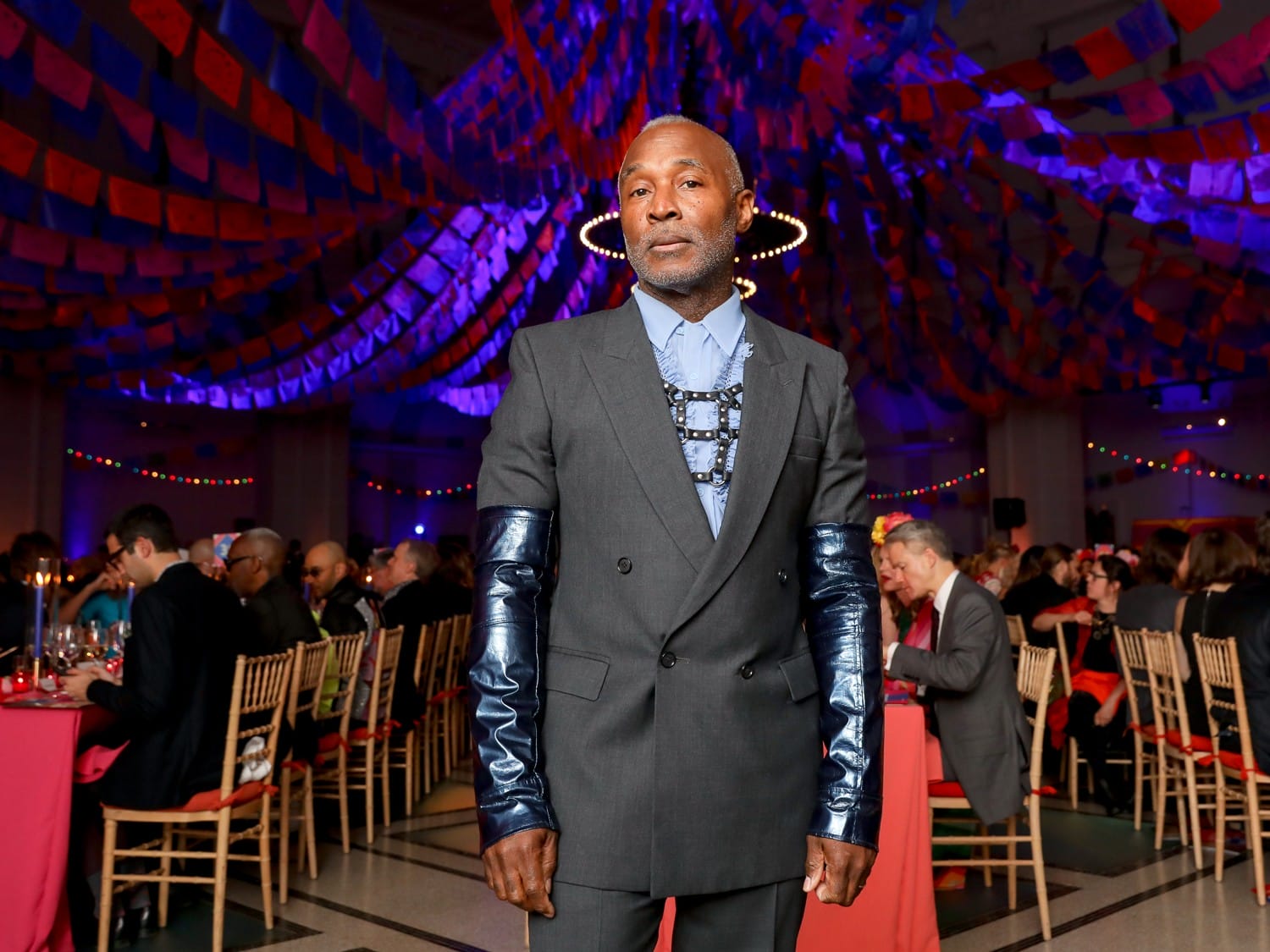 A person in a stylish dark suit with shiny sleeves standing in a vibrant, festive setting with colorful ceiling decorations.