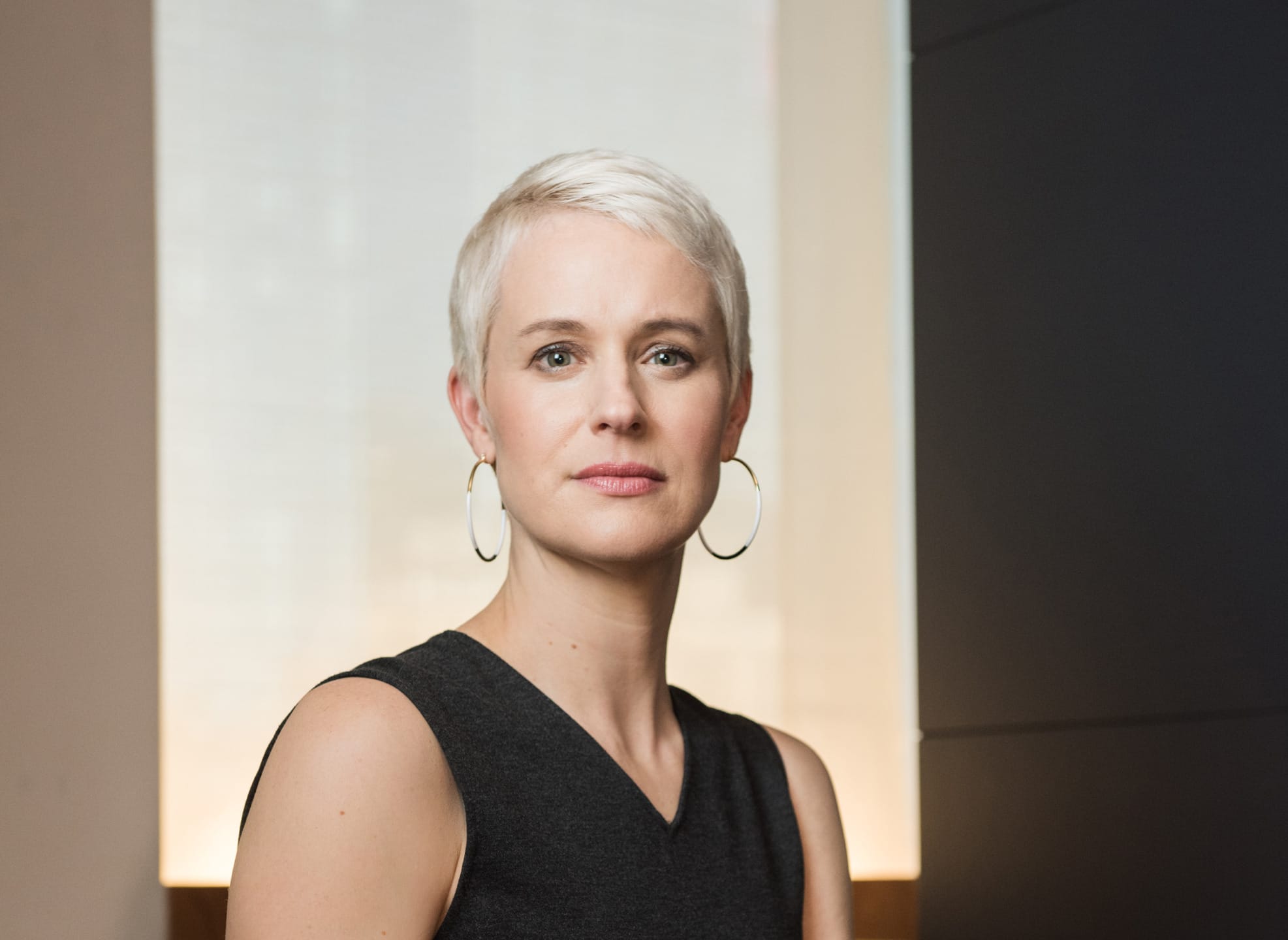 Person with short white hair wearing a black top and hoop earrings, standing in a softly lit indoor space.