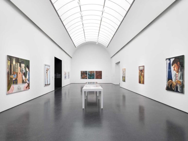 Modern art gallery with high ceiling, white walls, and paintings displayed along the walls.