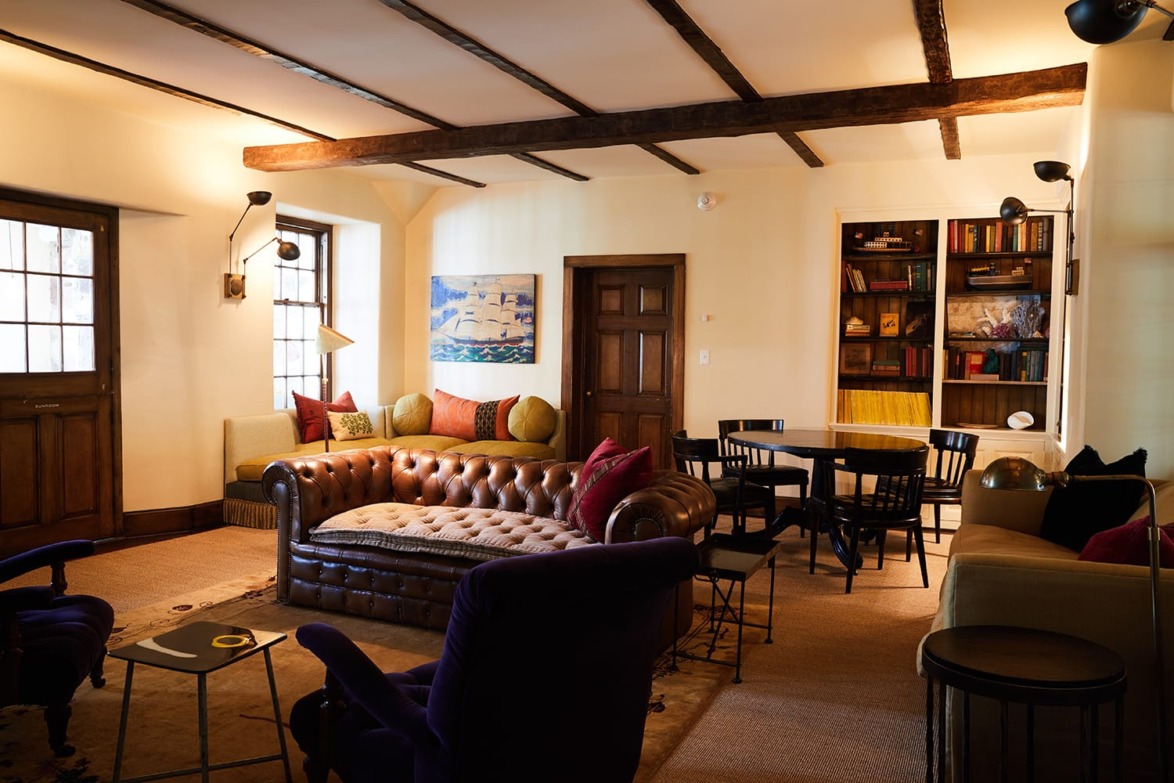 Cozy living room with a leather sofa, armchairs, bookshelf, artwork, and exposed wooden beams. Warm lighting creates a relaxed ambiance.
