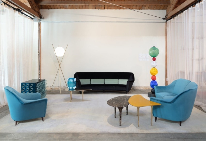 Mid-century modern living room with teal armchairs, black sofa, colorful lamp, and two small tables on a light rug.