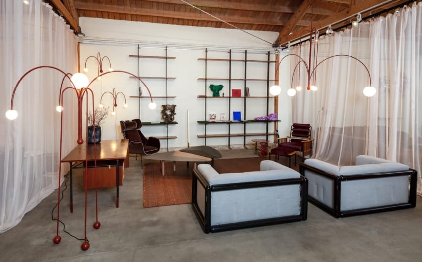 Modern office space with shelves, armchairs, unique lighting fixtures, and a wooden desk, enclosed by sheer white curtains.