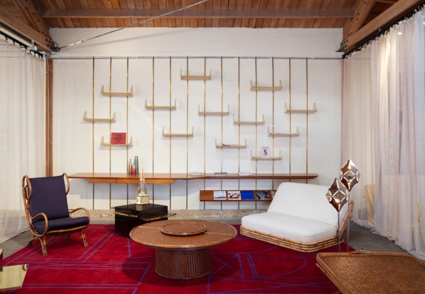 Modern living room with red geometric rug, rattan furniture, shelving with minimal decor, and wood ceiling beams.