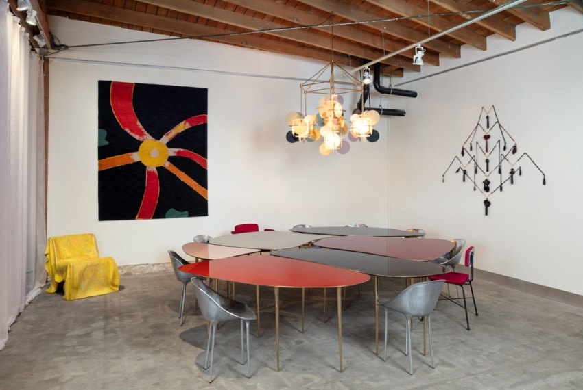 Modern conference room with triangular tables, colorful wall art, unique lighting fixture, and metal chairs on a concrete floor.