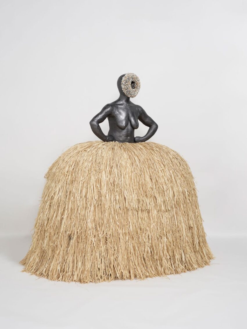 A figurative sculpture with a straw-like skirt and textured headpiece against a plain background.