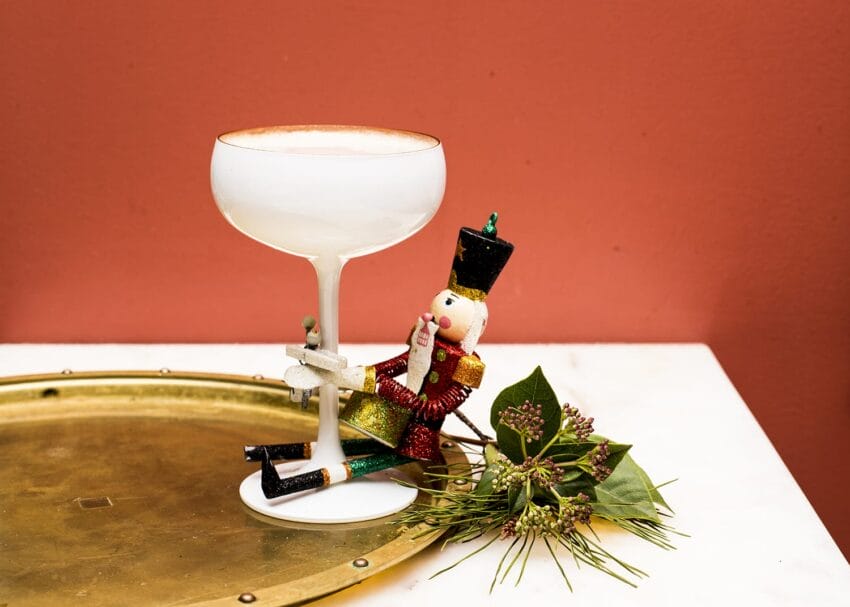 Nutcracker doll holding stemmed glass beside decorative greenery on brass tray with pink background.