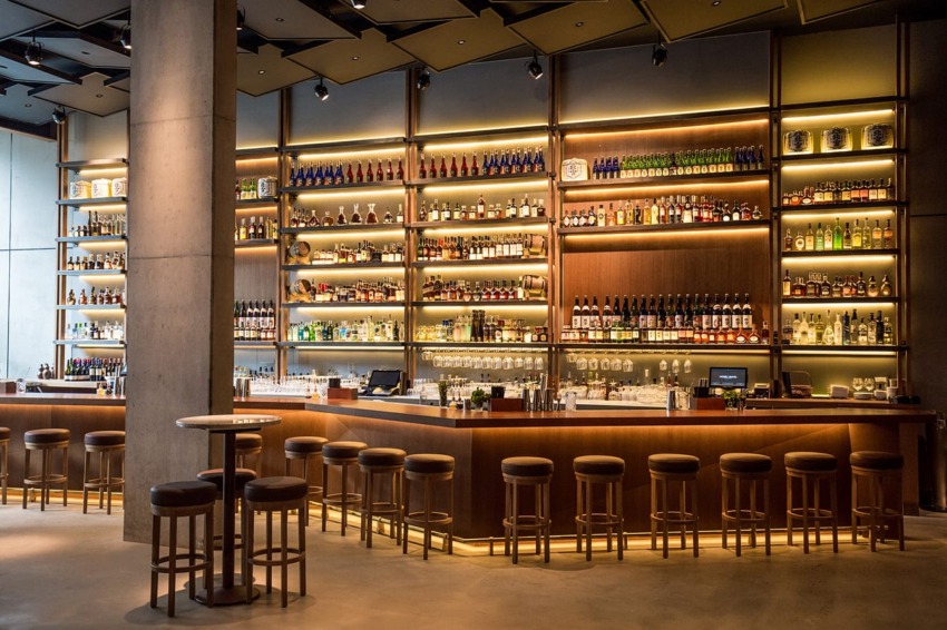 Modern bar interior with wooden shelves stocked with various bottles, a long counter, and high stools in a dimly lit setting.