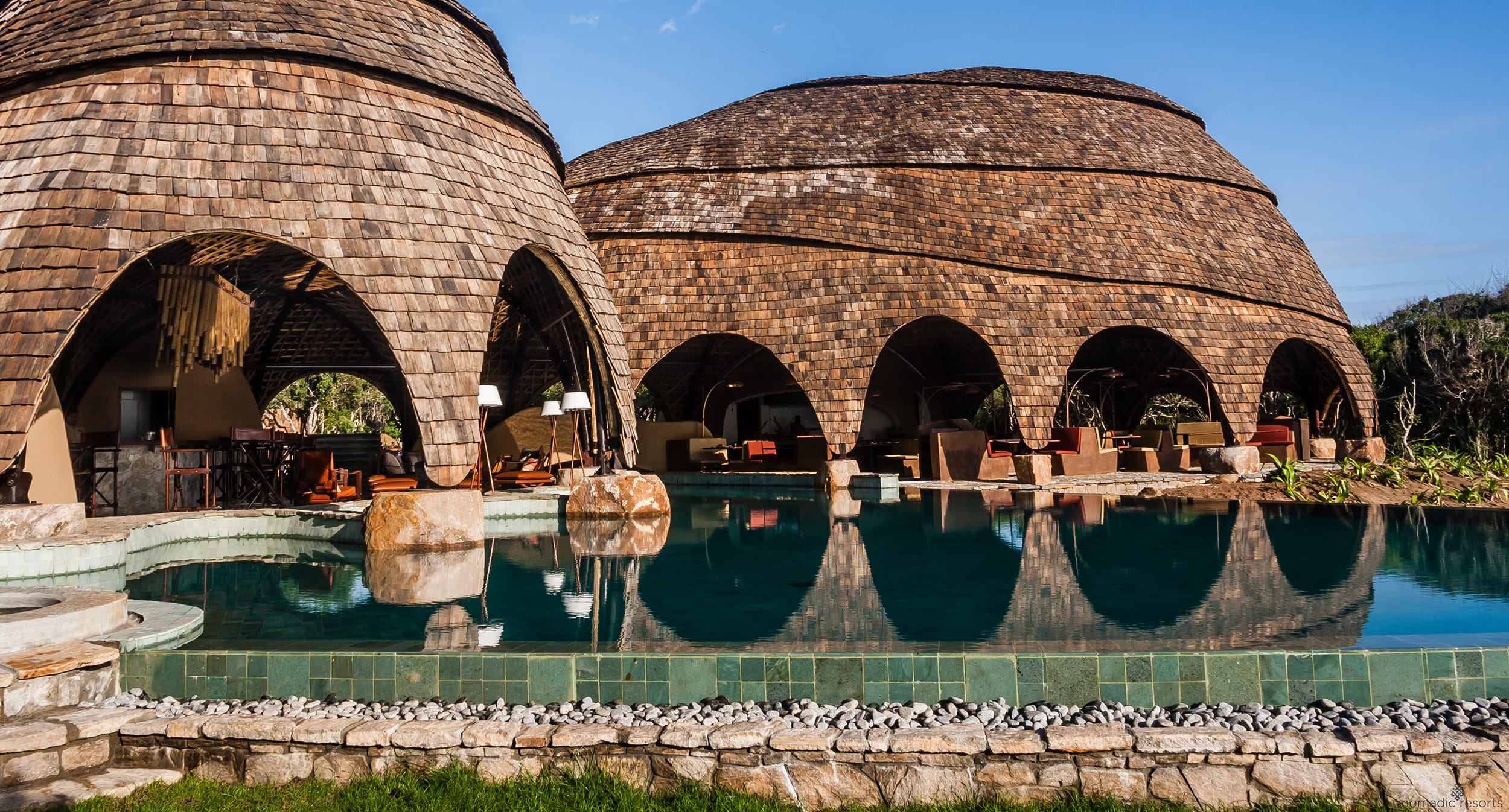 Resort with round stone structures and arched openings, reflecting in a clear blue pool surrounded by a natural landscape.