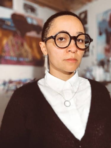 Person wearing glasses and a dark cardigan over a white shirt, standing in a room with blurry posters on the walls.